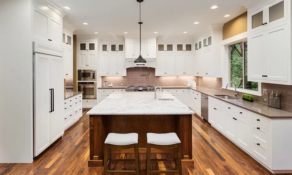 L-shaped modern kitchen with white shaker-style cabinets, a large wooden island with a marble countertop, and warm wooden flooring, featuring stainless steel appliances and elegant pendant lighting.