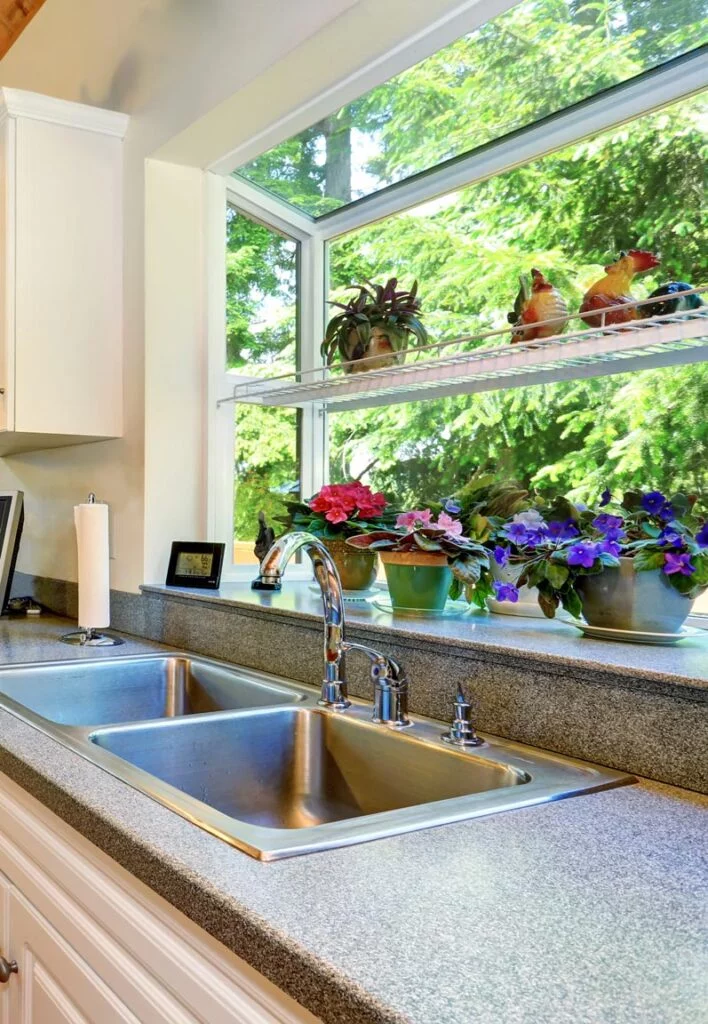 Bright L-shaped kitchen with a large garden window, double stainless steel sink, white cabinetry, and potted plants enhancing the natural ambiance.