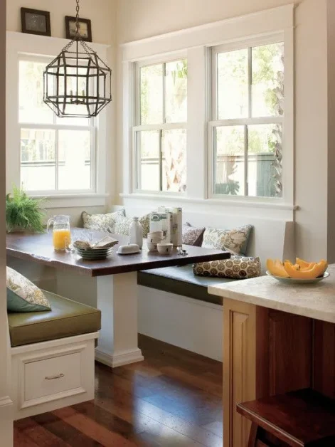 Modern L-shaped kitchen with a cozy built-in breakfast nook, white cabinetry, wooden flooring, large windows, and elegant pendant lighting.